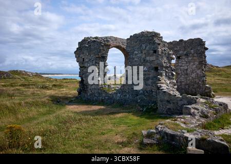 Die Ruinen der St. Dwynwens Kirche auf Llanddwyn Island vor Anglesey mit einem keltischen Kreuz im Hintergrund. Saint Dwynwen ist eine walisische Prinzessin aus dem 5. Jahrhundert Stockfoto