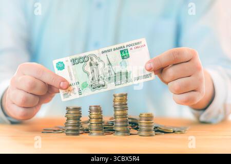 Geld sparen. Buchhaltungs-, Geschäfts-, Budget- und Finanzkonzept. Männliche Hände, die tausend russische Rubel-Banknoten über Münzstapeln halten Stockfoto