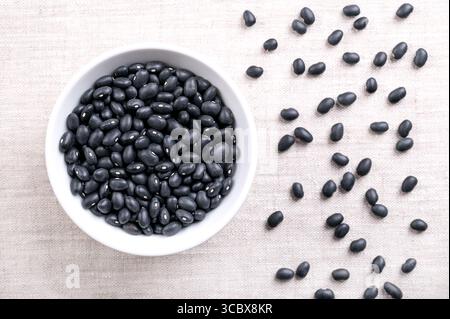 Schwarze Bohnen, in einer weißen Schüssel, auf Leinen. Getrocknete Sorte gewöhnlicher Bohnen, Phaseolus vulgaris, auch bekannt als schwarze Schildkrötenbohnen, und Kaviar criollo. Stockfoto