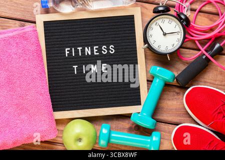 Fitnesszeit und Sportkonzept. Rote Turnschuhe, Apfel, Sprungseil, Kurzhanteln, rosa Handtuch, Flasche Wasser, Vintage-Wecker und schwarzes Schriftfeld mit Stockfoto
