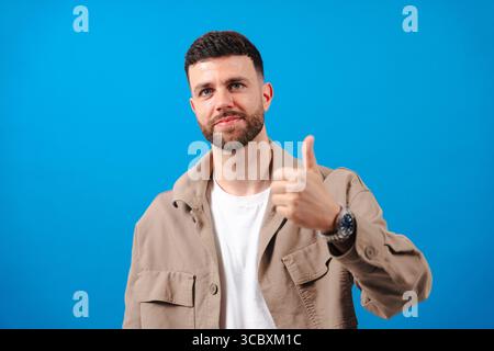 Selbstbewusster Mann in lässiger Jacke, der die Daumen nach oben zeigt, auf blauem Hintergrund Stockfoto