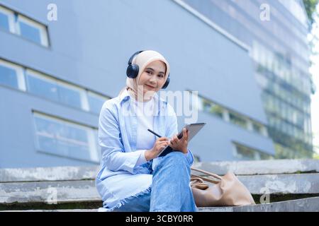Eine Frau mit Kopftuch sitzt auf einem Vorsprung mit Tablette und Stift. Sie schreibt oder macht Notizen. Konzept der Konzentration und Konzentration, Stockfoto