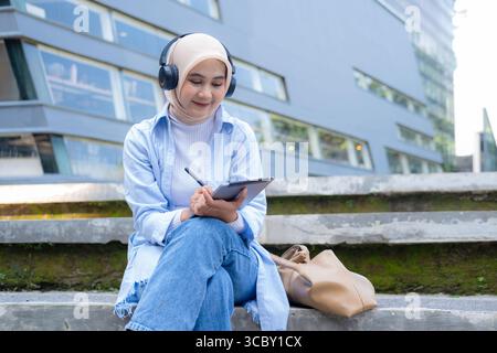 Eine Frau mit Kopftuch sitzt auf einer Bank mit einer Tablette im Schoß. Sie lächelt und genießt ihre Zeit Stockfoto
