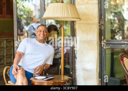 Ein lächelnder Mann sitzt beiläufig an einer Lampe in einem Café im Freien, sonnt sich im warmen Sonnenlicht und genießt einen entspannten Moment, umgeben von einer freundlichen Atmosphäre und Leuten, die sich in der Nähe unterhalten. Stockfoto