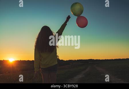 Mädchen, das bei Sonnenuntergang draußen mit bunten Ballons läuft. Kindheits- und Nachlässigkeitskonzept Stockfoto
