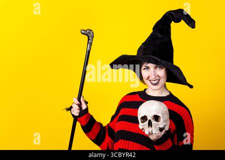 Halloween-Thema. Junge Hexe in gestreiftem Pullover, schwarzer Hut mit Schädel und Stock auf gelbem Hintergrund Stockfoto
