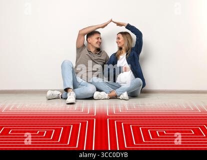 Schwangere Frau mit ihrem Mann, die mit ihren Händen Dach baut, während sie drinnen auf einem warmen Boden sitzt. Abbildung: Fußbodenheizung am Boden Stockfoto