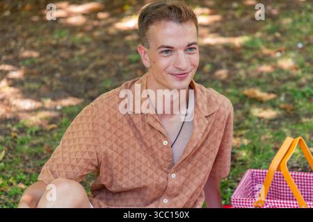 Porträt eines jungen Mannes, der beim Picknick in einem Park lächelt und bequem auf einer Decke sitzt, mit einem Picknickkorb gefüllt mit köstlichem Essen und Stockfoto
