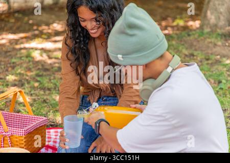 Junger Mann, der frischen Orangensaft in ein Glas gießt, das von einer lächelnden jungen Frau gehalten wird, während er an einem sonnigen Tag im Park ein herrliches Picknick genießt Stockfoto