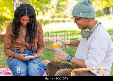 Multikulturelle Freunde genießen ein sonniges Picknick im Park und teilen Orangensaft und Snacks, umgeben von lebendiger Natur und füllen die Luft mit Stockfoto