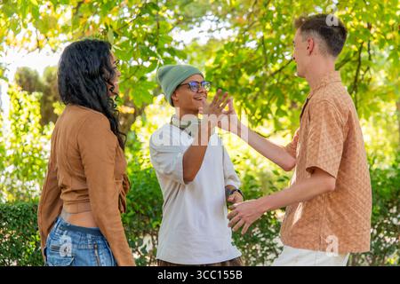 Drei junge Freunde, eine Frau und zwei Männer, genießen ein Picknick in einem Park, lachen und Gesten, während sie gemeinsam einen glücklichen Moment verbringen, umgeben von Stockfoto