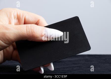 Weibliche Hand mit weißer Maniküre hält die schwarze Bankkarte in Nahaufnahme gegen eine helle Wand Stockfoto