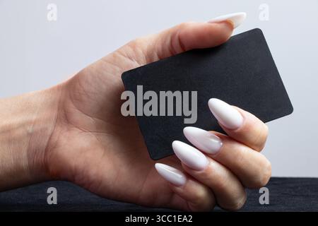 Weibliche Hand mit weißer Maniküre hält die schwarze Bankkarte in Nahaufnahme gegen eine helle Wand Stockfoto