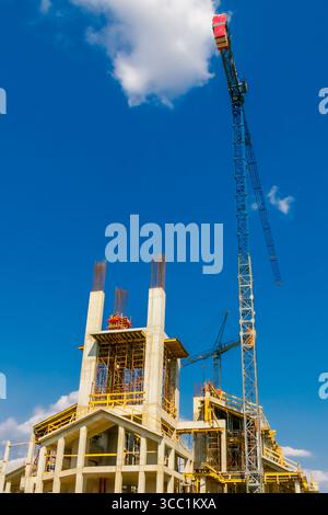 Arbeiter sind auf einer Baustelle beschäftigt, auf der ein großer Kran Materialien hebt. Eine sich entwickelnde Gebäudestruktur zeigt einen Betonrahmen, der bis zum Schlepptau reicht Stockfoto