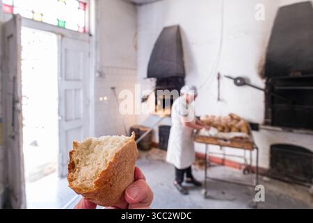 Hand hält Favaios Brot, während Bäckerin im Hintergrund arbeitet in Rosalia Bakery, Favaios, Alijó, Vila Real District, Nordregion, Portugal Stockfoto