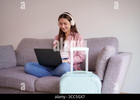 Fröhliche asiatische Frau benutzt Computer und Kopfhörer auf einem Sofa neben dem Gepäck. Sie bereitet sich auf eine Reise vor oder arbeitet aus der Ferne. Stockfoto