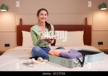 Erwachsene asiatische Frau plant nachdenklich ihre aufregende Urlaubsreise in einem gemütlichen Hotelzimmer. Sie bereitet das wesentliche für eine unvergessliche Reise vor. Stockfoto