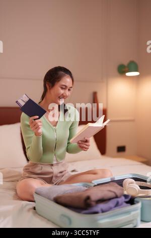 Happy Asian Woman bereitet Reise vor Lesen Reiseführer arrangiert Koffer bereit für aufregende Urlaubsabenteuer Stockfoto