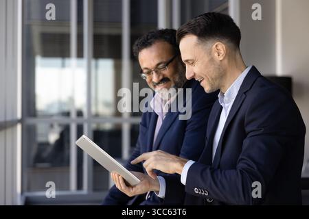 Zwei glückliche, vielseitige jüngere und ältere Geschäftskollegen, die ein Tablet verwenden Stockfoto