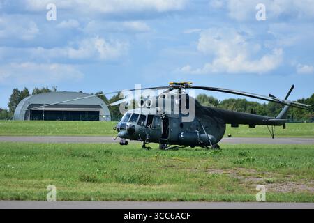 Großer Mi-17 Militärtransporthubschrauber auf grasbewachsenem Flugplatz, Vorderansicht mit ausgefahrenen Rotorblättern, Cockpitfenster reflektieren Sonnenlicht, Hangar in BA Stockfoto