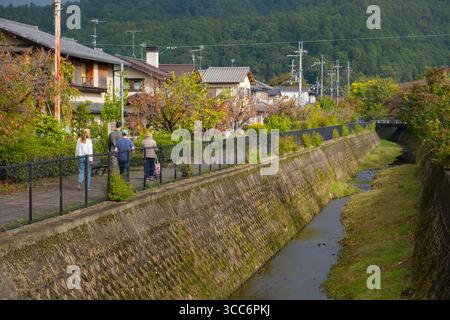 Eine Familie genießt einen gemütlichen Spaziergang entlang eines von Bäumen gesäumten Pfades neben einem kleinen Fluss in einer ruhigen Wohngegend, die die Essenz der Entspannung und der Entspannung einfängt Stockfoto