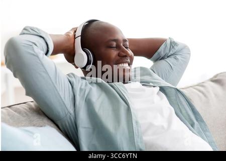 Fröhlicher, schwarzer, reifer Mann mit schnurlosem Headset, der sich auf dem Sofa entspannt und lächelt, während er mit geschlossenen Augen Musik hört Stockfoto