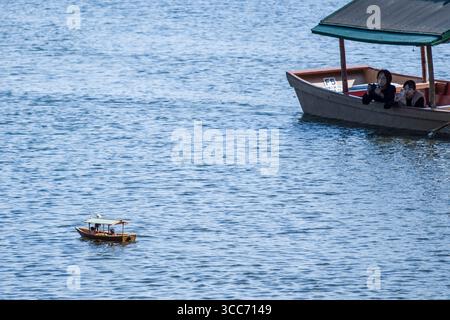 Eine friedliche Szene, in der zwei Personen in einem Boot ein kleines hölzernes Modellboot beobachten, das auf einem ruhigen See schwimmt. Das ruhige Wasser reflektiert die Ruhe A Stockfoto