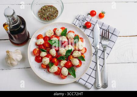 Italienischer Caprese-Salat mit Kirschtomaten, Mozzarella, Basilikum, Balsamico-Sauce auf weißem Holztisch. Draufsicht, flach. Stockfoto