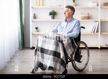 Trauriger behinderter älterer Mann, der allein im Altersheim auf dem Rollstuhl sitzt und durch das Fenster schaut Stockfoto