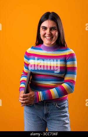 Eine junge Frau mit einem einzigartigen Stil lächelt selbstbewusst und hält ein Tablet. Sie steht vor einem leuchtend orangefarbenen Hintergrund und zeigt ihre lebendige Person Stockfoto