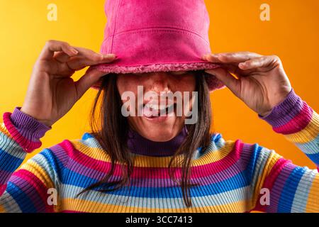 Die junge Frau zeigt ihren einzigartigen Stil mit einem verspielten Ausdruck. Sie trägt ein lebhaftes Outfit und einen rosa Hut vor einem auffälligen orangefarbenen Hintergrund, um eine se festzuhalten Stockfoto