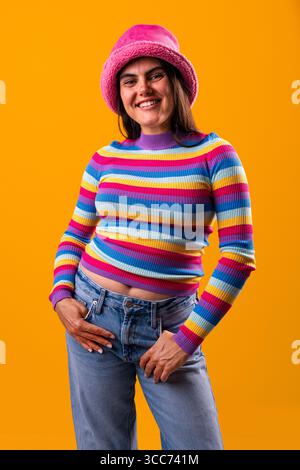 Die junge Frau zeigt ihren einzigartigen Stil mit einem lebendigen, farbenfrohen Outfit und einer selbstbewussten Pose vor orangefarbenem Hintergrund, die ihre Individualität unterstreicht Stockfoto