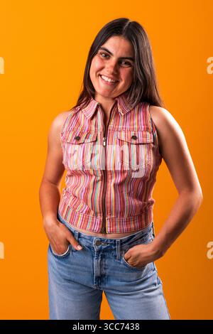 Fröhliche junge Frau mit einzigartigem Stil, mit einem farbenfrohen gestreiften ärmellosen Oberteil und einer Jeans, die selbstbewusst mit Händen in Taschen gegen eine Aue posiert Stockfoto