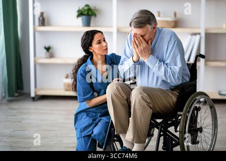 Depressive ältere Männer im Rollstuhl weinend, junge Krankenschwester tröstet ihn im Altersheim Stockfoto