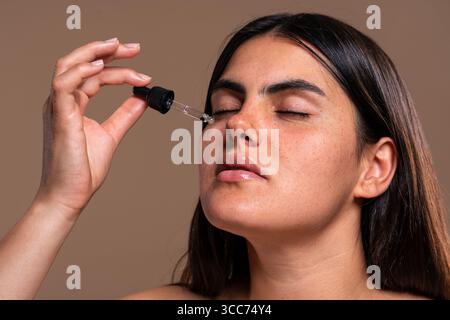 Die schöne junge Frau mit Sommersprossen schließt ihre Augen und trägt mit einem Tropfer ein feuchtigkeitsspendendes Serum auf ihr Gesicht auf, was die Gesichtspflege, Körperpflege, an fördert Stockfoto