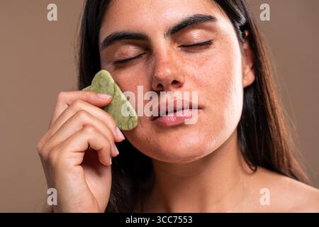Junge Frau mit geschlossenen Augen mit einem Jade Gua Sha Schaben Massagewerkzeug auf ihrem Gesicht, fördert Lymphdrainage, reduziert Schwellungen und verbessert c Stockfoto