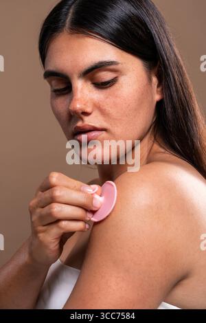 Konzentrierte junge Frau mit Sommersprossen, die ihre Schultern mit einer rosa Silikonbürste peelt und Hautpflege und Körperpflegeroutinen für gesunde, Stockfoto