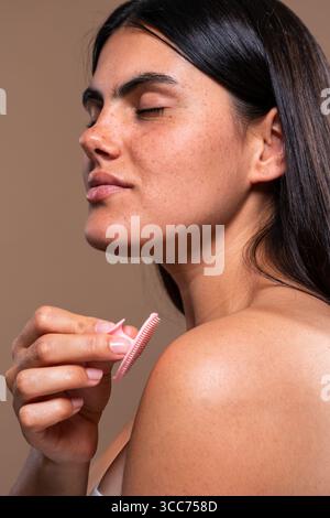Mit geschlossenen Augen und einem ruhigen Ausdruck peelt eine junge Frau sanft die Haut ihrer Schulter mit einer rosafarbenen Silikonbürste und nimmt einen Moment von ein Stockfoto