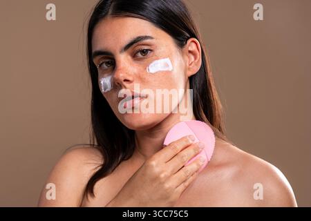 Schöne junge Frau mit Sommersprossen trägt Feuchtigkeitscreme auf ihr Gesicht auf und verwendet eine rosa Silikon Gesichtsreinigungsbürste für eine vollständige Hautpflege Stockfoto