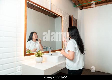 Asiatische Frau, die vor einem Badezimmerspiegel steht und Lippenstift aufträgt. Selbstpflege- und Schönheitsroutine am Morgen. Stockfoto