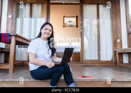 Eine junge indonesische Frau lächelt, während sie an einem Laptop vor einer gemütlichen Holzhütte arbeitet. Perfekt für Lifestyle, Fernarbeit, Freiberufler und Reisen Stockfoto