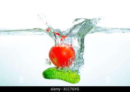 Dynamischer Spritzer einer frischen roten Tomate in klares Wasser mit einer Gurke, die Frische und Vitalität vor einem hellen Hintergrund unterstreicht. Stockfoto
