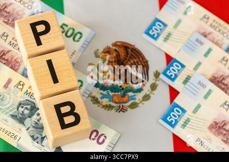 Wirtschaft in Mexiko. Das Wort PIB (Bruttoinlandsprodukt) steht auf Holzwürfeln, mit mexikanischem Geld in gelber mexikanischer Flagge. Nahaufnahme Stockfoto