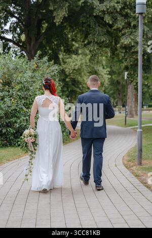 Braut und Bräutigam gehen Hand in Hand auf einem geschwungenen Parkweg, umgeben von üppigen grünen Bäumen. Die Braut hält einen kaskadierenden Rosenstrauß Stockfoto