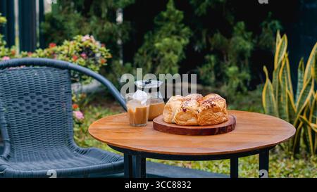 Frisches Brot und Eiskaffee werden auf einem runden Holztisch im Freien serviert, mit verschwommenem Grün im Hintergrund. Stockfoto
