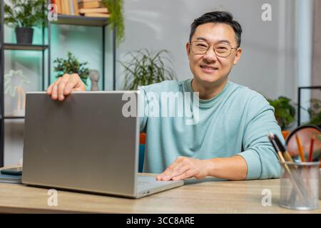 Asiatischer reifer Mann Freiberufler zu Hause Wohnzimmer Wohnung und sitzend auf Couch am Tisch Schreibtisch, öffnet Laptop. Ein Chinese, der an einem Notebook arbeitet, Nachrichten sendet, Online-Einkäufe tätigt, Filme ansieht Stockfoto