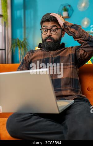 Verärgerter junger Indianer, der zu Hause auf dem Sofa sitzt und einen Laptop benutzt, der Angst und Stress hat. Schockierter Araber, der auf Verlust reagiert, schlechte Nachrichten über Netbook in Wohnzimmerwohnung. Computervirus. Vertikal Stockfoto
