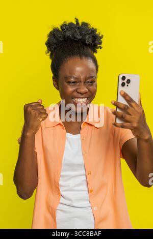 Glücklich aufgeregt afrikanische Schwarze Frau Mädchen verwenden Smartphone Tippen surfen schreien sagen Wow Ja fand heraus, großer Gewinn gute Nachrichten Lotterie Zielerreichung feiert Erfolg, gewinnt Spiel auf gelb Stockfoto