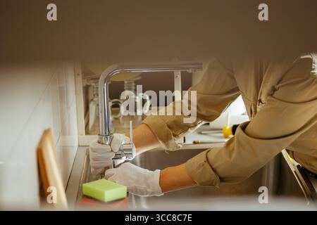 Ein Klempner repariert einen Chromhahn in einem Küchenspüle mit Handschuhen. Die Szene zeigt die fachkundige Hausreparatur und den Schwerpunkt auf die Küchenpflege. Stockfoto