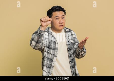 Klopfen-klopfen, wer da ist. Verwirrter aufgeregter asiatischer Mann, der an die Tür klopft und fragt, wer zu Hause ist, sich peinlich fühlt, keine Ahnung hat, ahnungslos und unsicher ist. Reifer Chinese auf beigefarbenem Hintergrund Stockfoto